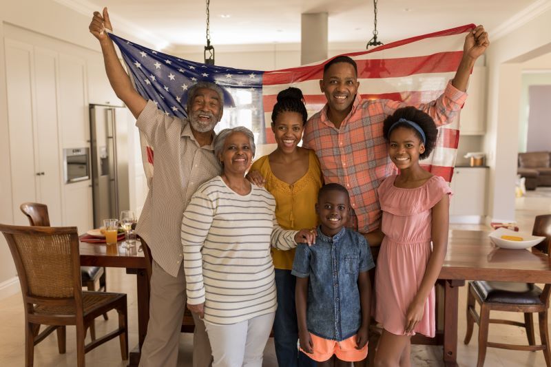 A multi-generational family smiling together in their home, with an elder man holding up an American flag, symbolizing their immigrant heritage. The family, dressed in casual attire, represents three generations, including grandparents, parents, and two young children, radiating happiness and unity.