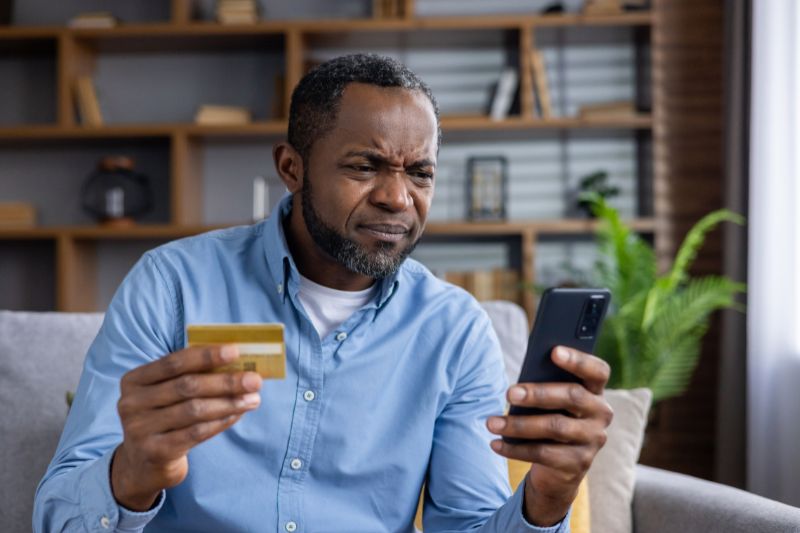 Consumer reviewing credit card and phone looking concerned about credit repair company, representing CROA violations, credit repair company sued, and legal action for credit repair law violations
